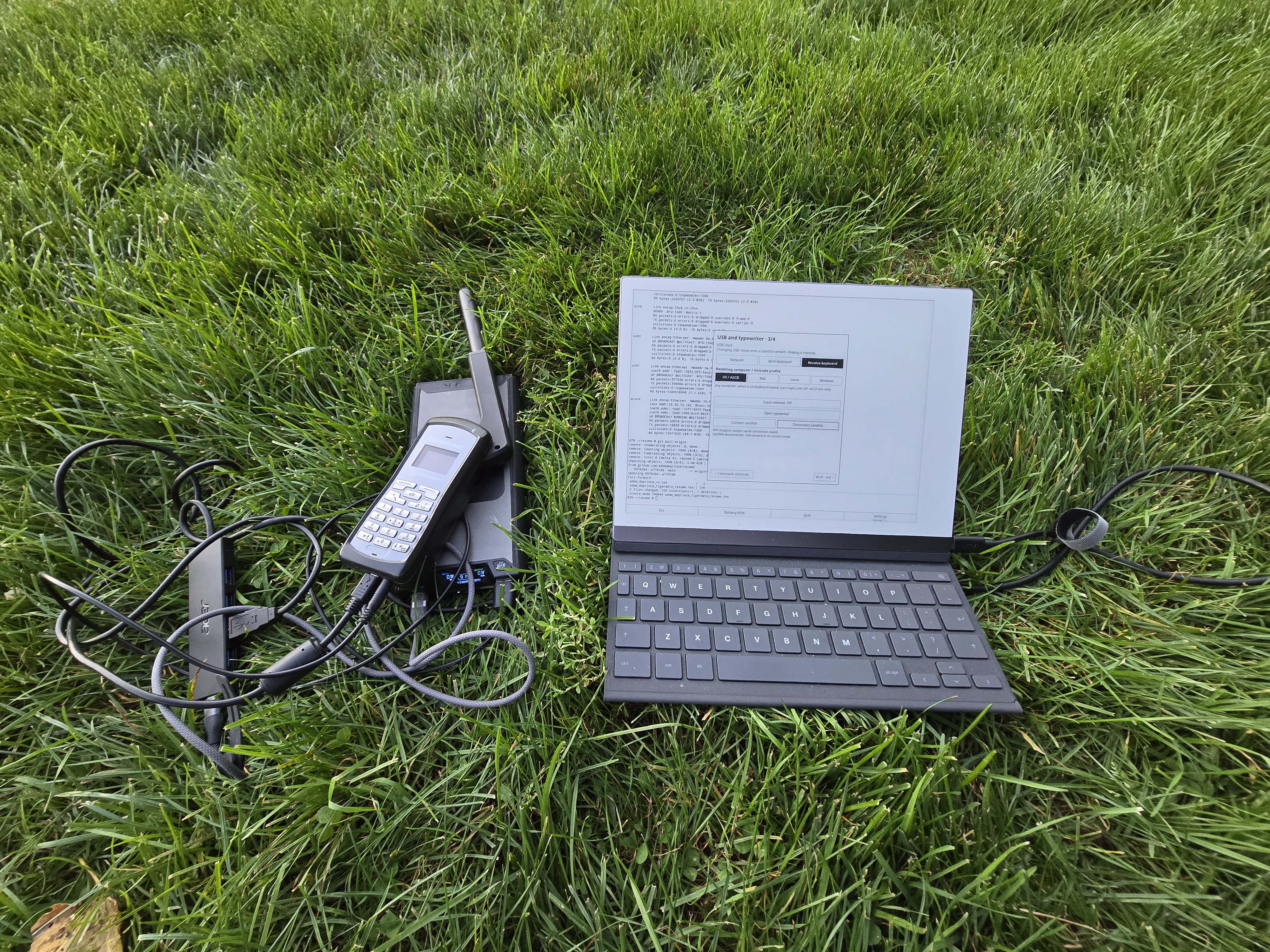 A reMarkable 2 with a keyboard running Inkline on the grass beside a GSP-1700 satellite phone, power bank, powered USB hub, and connecting cables.